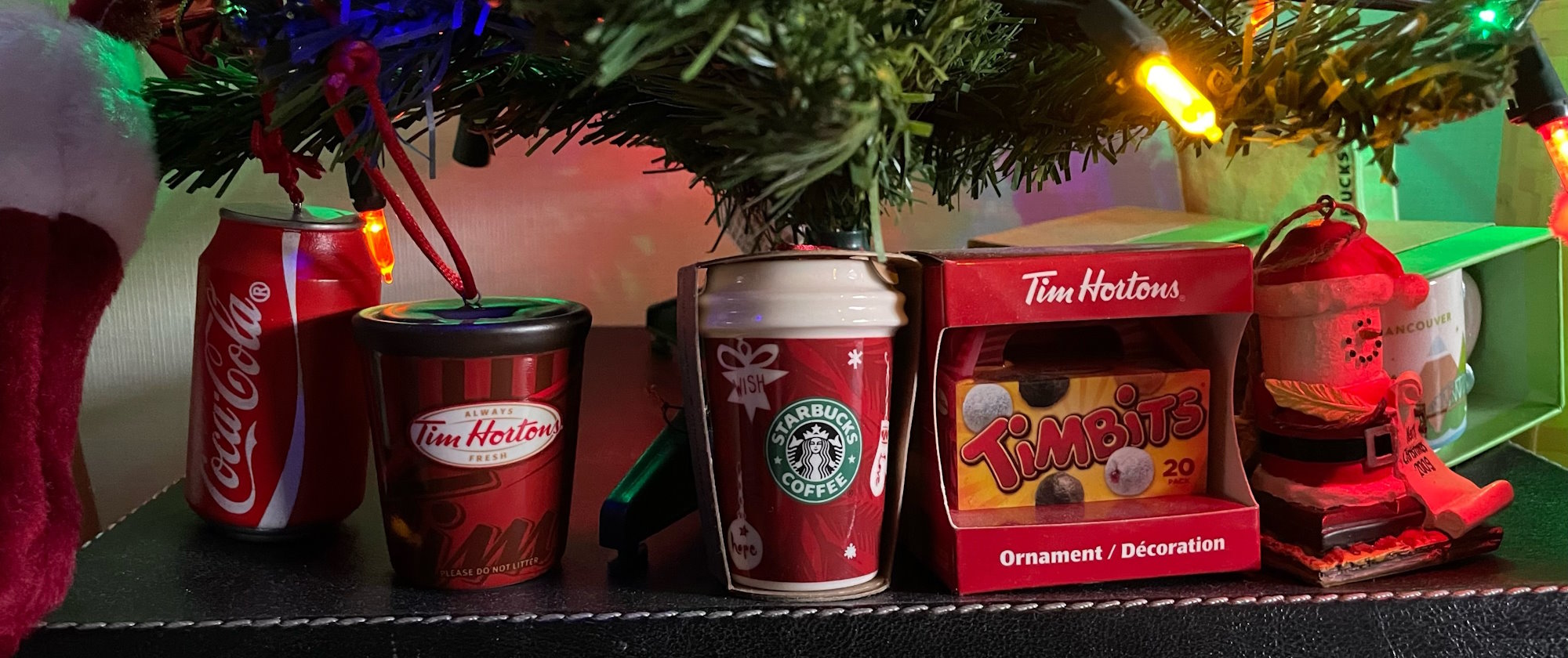 The bottom of a small plastic xmas tree standing on a black faux leather banker’s box. A row of ornaments are arrayed beneath the tree like presents: a Coca-Cola can, a Tim Hortons coffee cup, a Starbucks cup, a Timbits box, a marshmallow dressed like Santa sitting on a graham cracker, a Starbucks mug that says Vancouver.