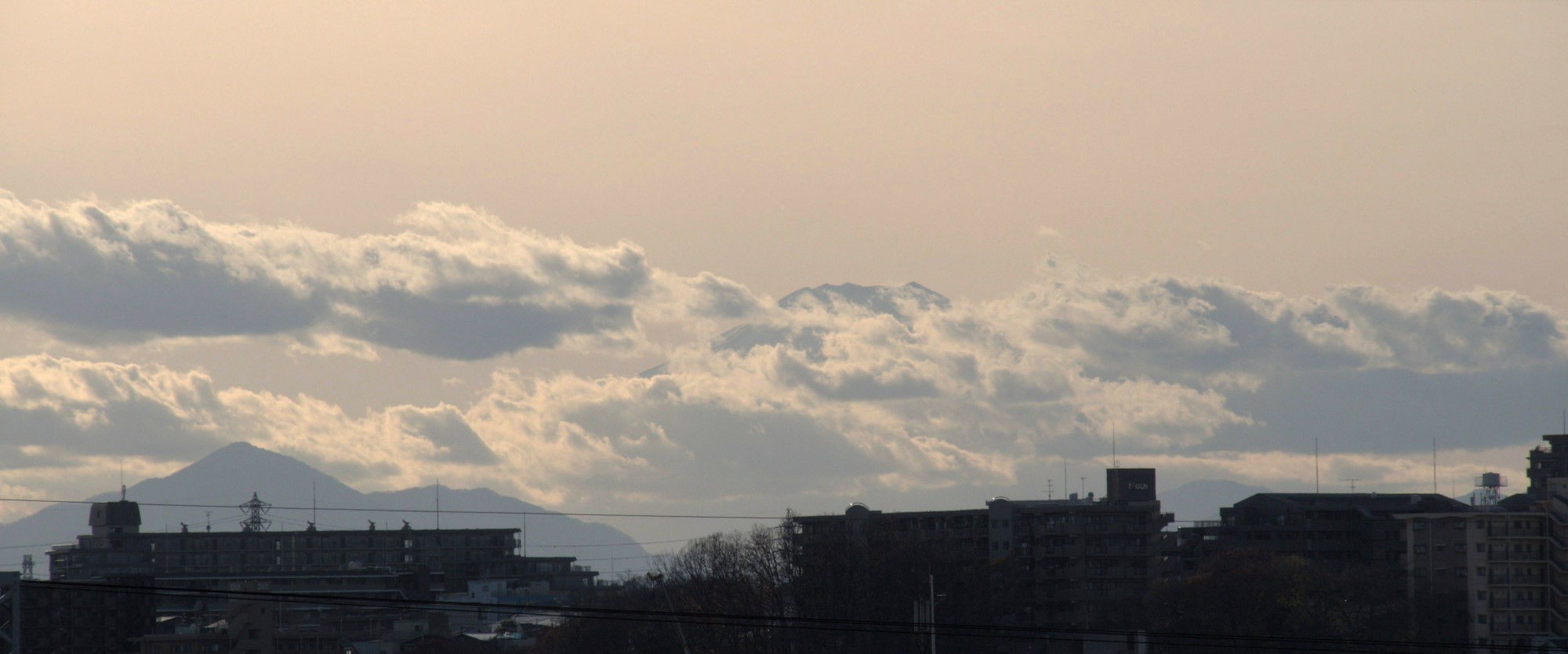 A wide telephoto shot of Mount Fuji rising just barely above a band of clouds beyond some neighbourhood apartment buildings in dark silhouette. The mountain, sky, and clouds are all a light sandy colour through the haze and strong back-lighting by the sun.