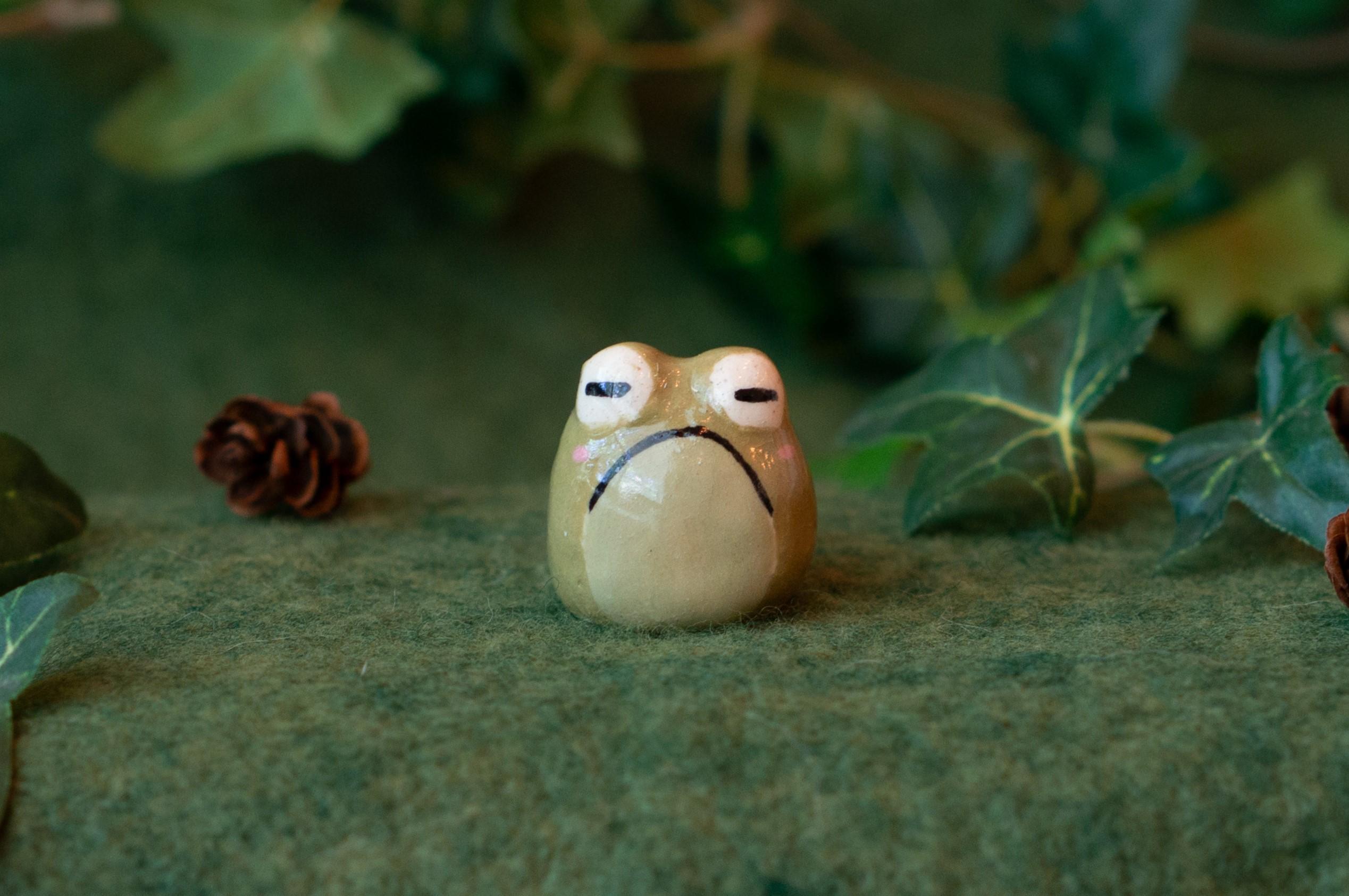 A tiny ceramic toad figurine with a cute, grumpy expression. The figurine is rounded, about the size of a large cherry tomato, with bulging eyes and no limbs. The body is a light olive color, and is smooth and glossy. Its face is hand-painted and consists of large white eyes with horizontal slit pupils, a large downturned mouth, and pink cheeks. The backside (not shown in this picture) is bumpy as if it’s covered in warts. The figurine is set on a green felt backdrop with decorative faux greenery (ivy) and tiny pinecones.