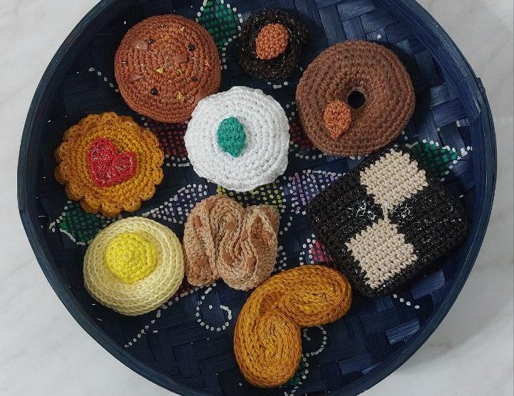 On a white surface: a blue woven basket filled with various crochet biscuits including a donut shaped biscuit with an almond on top, a Jammie Dodger, a lemon biscuit, a French palmier, an Italian cassatina, and other brown and beige biscuits with decorations on top.
