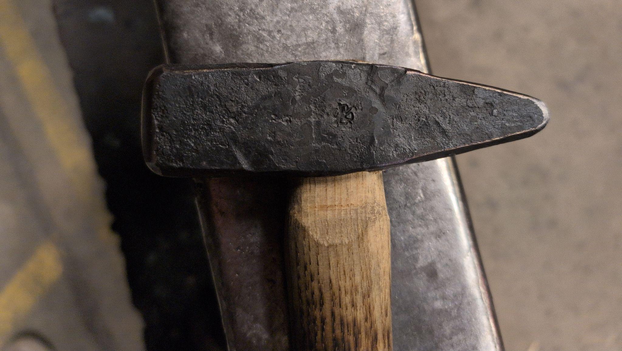 A close up of the head of a cross peen hammer sitting on an anvil. The wood  handle is blackened by fire and has been lightly oiled. The hammer head is blackened, typical of forged steel and iron. In the middle of the hammer head, there is an impression of a fireweed flower, the maker&rsquo;s mark. 