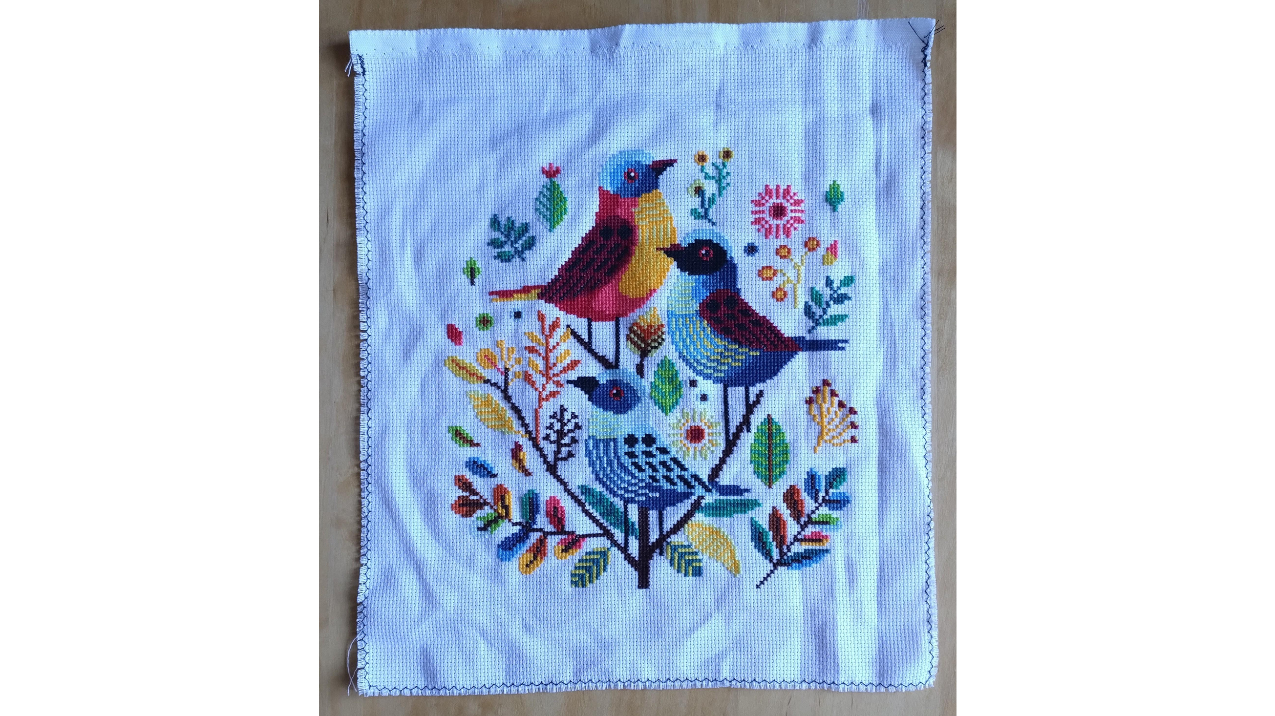 Finished cross stitch pattern with three colourful birds in the middle, surrounded by leaves.
